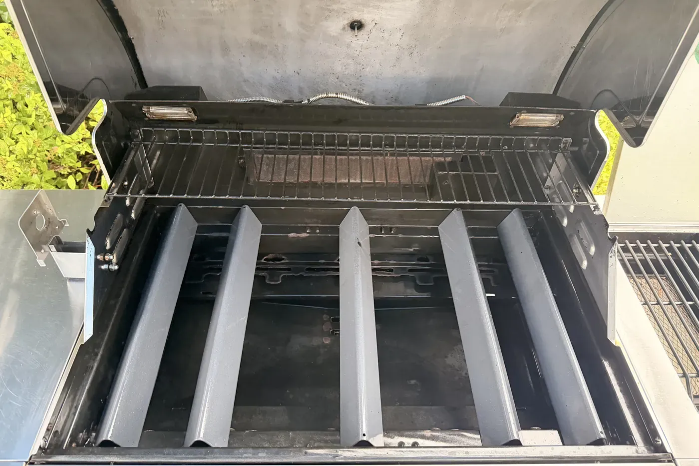Inside a built-in gas grill after a restoration, lid open, with five V-shaped heat shields over the burners, a clean warming rack and the rear rotisserie burner visible
