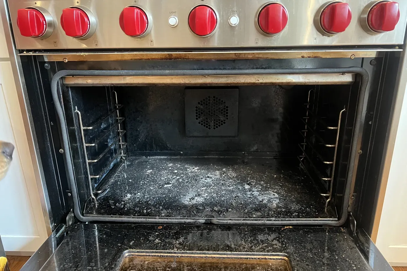 Pro range oven with red knobs and the door open before a cleaning, the cavity floor covered in gray ash and carbon flakes, a dark gasket running around the opening and a round convection fan cover on the back wall