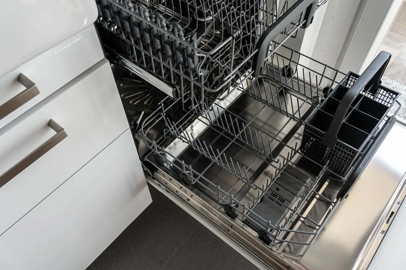 Stock photo of an open stainless dishwasher with its empty racks and cutlery basket pulled out in a white kitchen