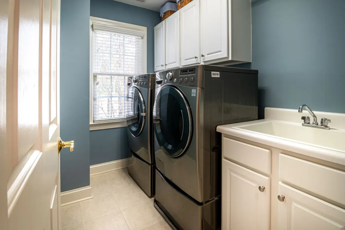 Stock photo of a laundry room with a front-load washer and dryer on pedestals, white cabinets above and a utility sink beside them