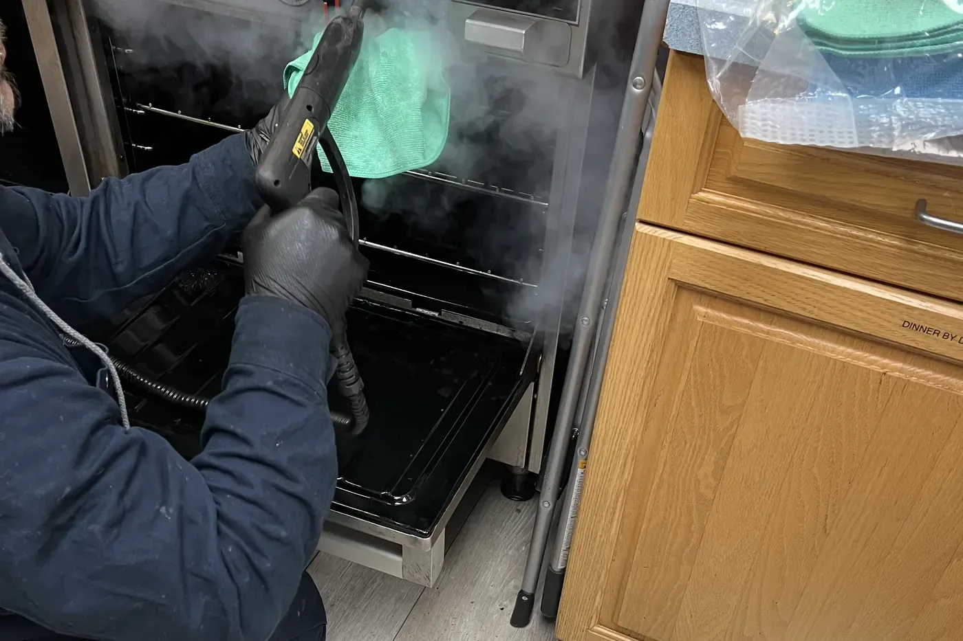 A technician in black gloves steam-cleaning the inside of an oven with a steam wand and a green microfiber cloth, plastic sheeting on the floor beside him
