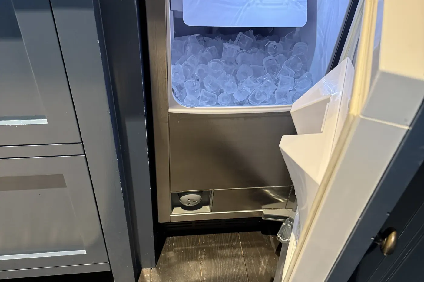 Sub-Zero under-counter ice maker with its door open and the bin full of clear ice, set into navy cabinetry under a dark granite counter
