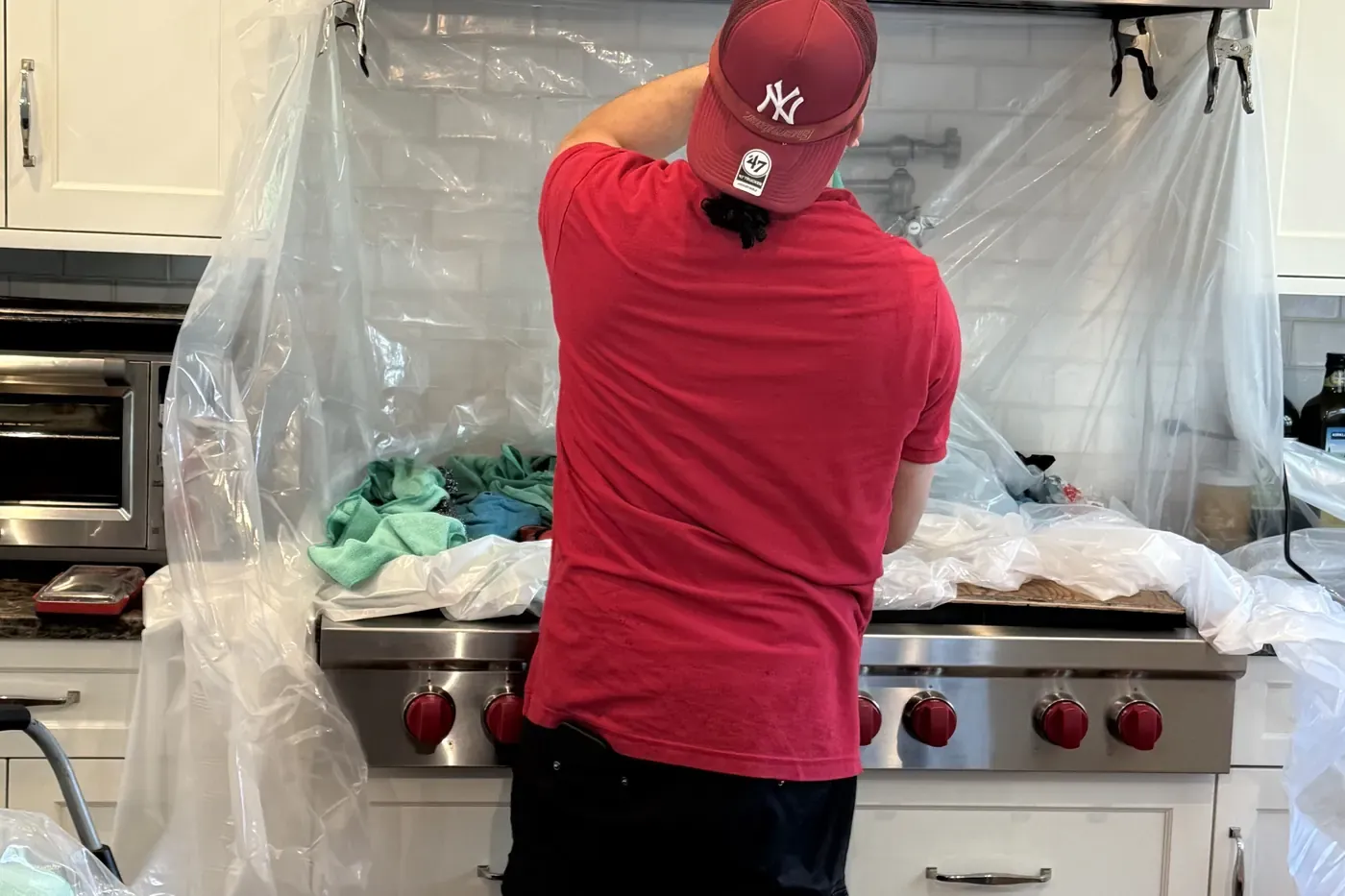 One of our technicians working at a Wolf range with red knobs under a stainless chimney hood, plastic sheeting clamped to the hood edge and draped over the backsplash, counters and cabinets