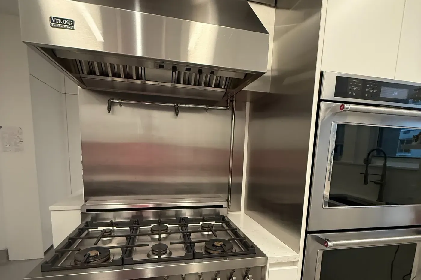 Viking Professional wall hood with baffle filters over a Bertazzoni gas range and a stainless backsplash with a pot filler, KitchenAid double wall oven beside it, white cabinets