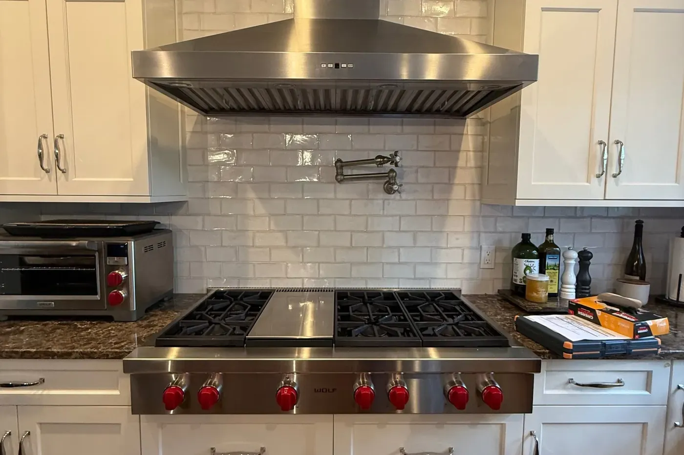 Stainless chimney range hood with baffle filters hanging over a six-burner Wolf rangetop with red knobs and a center griddle, in a white kitchen with glazed subway tile, a brass pot filler and dark granite counters