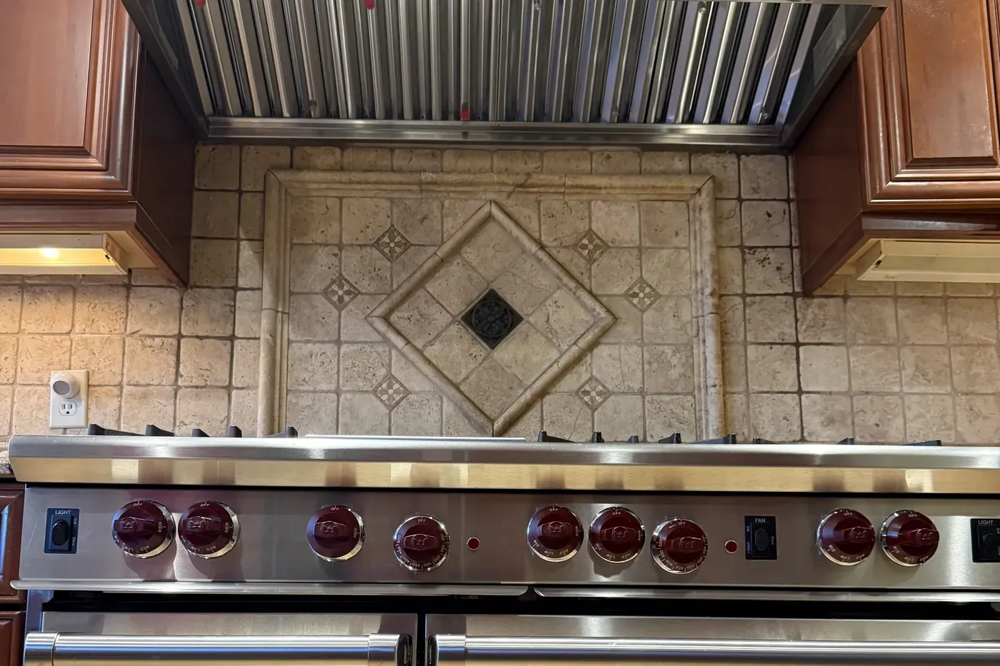 Wolf 48-inch range with red knobs under a stainless pro hood liner with baffle filters and lights, tumbled stone backsplash with a diamond inset, cherry cabinets